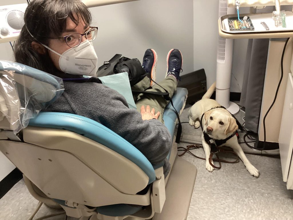 Woman with dark hair, glasses, and a white mask in a dentist chair with a yellow Lab service dog lying at the foot of the dentist chair, under the instrument tray.