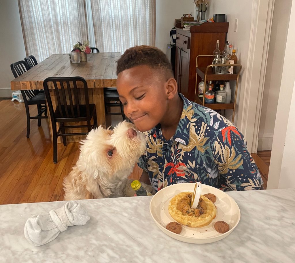 Fluffy Labradoodle leans up to kiss the chin of a boy wearing a Hawaain shirt, with a waffle with a candle in the shape of a 9 on the counter in front of him.