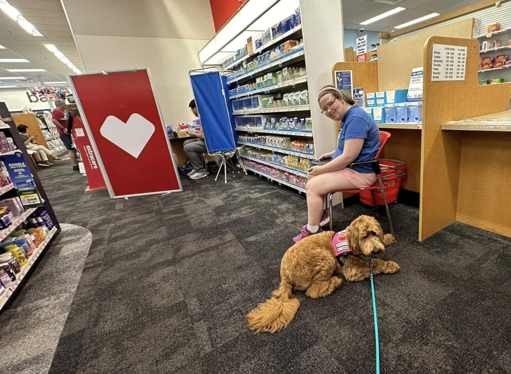 Kati sits on chair at CVS pharmacy counter while Heidi lies on the floor in front of her.