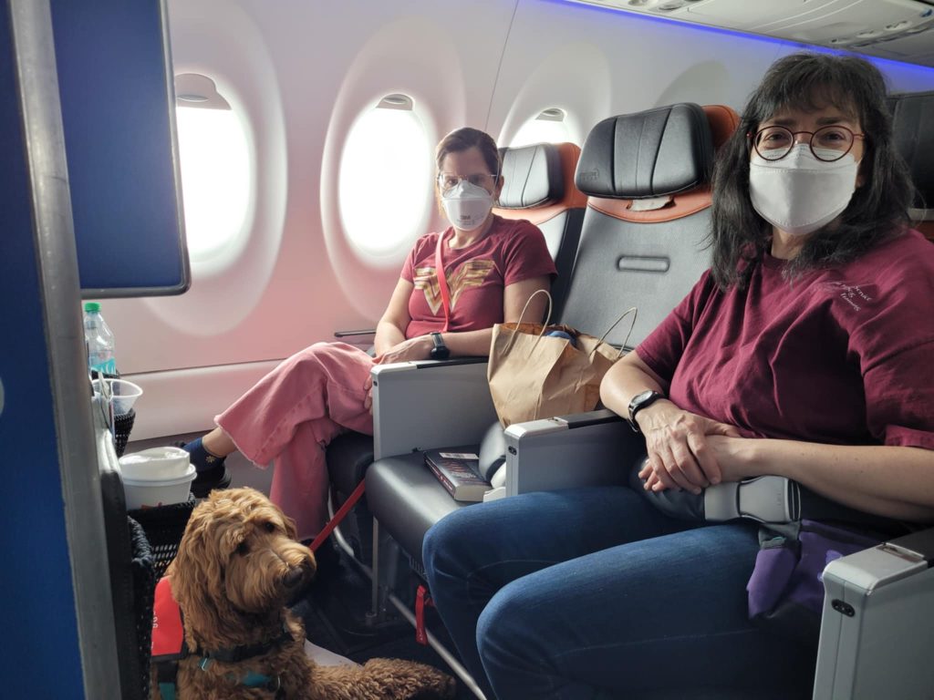 Tere sits next to the window seat in an airplane with Heidi on the floor in front of her and Sharon in the aisle seat.